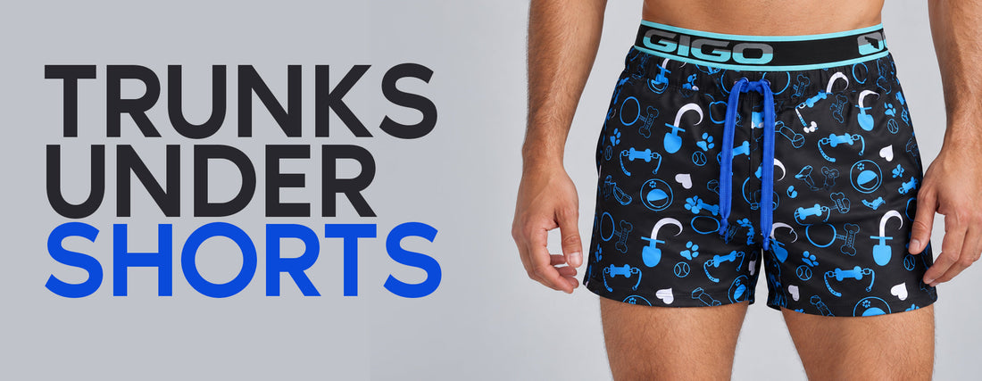 Model wearing black shorts with blue-and-white prints over Gigo trunks with a visible waistband, beside the text “Trunks Under Shorts”.